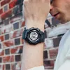 A person wearing a black digital watch on their wrist, with a brick wall in the background. Produktbild-Vorschau