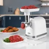 A white meat grinder is shown in a kitchen setting, with raw meat in the hopper and ground meat on a plate. Anteprima dell’immagine prodotto