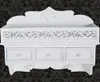 A white decorative wall-mounted shelf with three drawers and two hooks against a dark patterned background. Produktbild-Vorschau