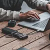 A person's hands are using a laptop on a wooden table, with a card reader and a camera nearby. Produktbild-Vorschau