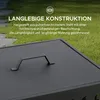 A close-up view of a black metal fire pit lid with a handle, set against a blurred background of green grass and stone paving. Náhled obrázku produktu