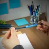 Hands holding a blue pen over a desk with other pens in a jar and sticky notes on the wall. Podgląd zdjęcia produktu