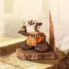 A figurine of the robot Wall-E holding a plant in a boot, standing on a rocky base with a tall, abstract structure behind it. The background suggests a hazy, urban environment. Anteprima dell’immagine prodotto