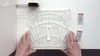 Hands holding a clear plastic template on a white surface, with a measuring tape and two small red and silver packets nearby. Anteprima dell’immagine prodotto