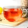 A clear glass mug filled with a red beverage sits on a wooden board, next to a lemon and some tea leaves. In the background, there are jars of dry ingredients. Voir les images du produit