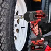 A hand wearing a black glove is holding a red and black cordless impact wrench, using it to tighten a lug nut on a car wheel. Voir les images du produit