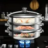 A multi-tiered stainless steel steamer pot is shown on a stovetop with a blue flame underneath. The different tiers are cut away to reveal food being steamed: dumplings on the top, shrimp in the middle, and fish on the bottom. Arrows indicate steam circula Produktbild-Vorschau