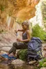 A woman is sitting by a stream in a canyon, opening a bottle. A large backpack is next to her. Podgląd zdjęcia produktu