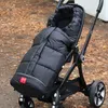 A black stroller with a puffy black footmuff is parked outdoors on a surface covered with fallen leaves. Produktbild-Vorschau