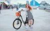 A woman with red hair is posing with an electric bicycle on a paved walkway near a body of water. There are buildings and other people in the background. Voir les images du produit