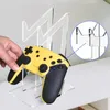A hand is placing a yellow and black game controller onto a clear acrylic stand. Produktbild-Vorschau