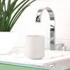 A soft grey ceramic toothbrush holder sits on a white countertop next to a modern chrome faucet and a potted plant. Produktbild-Vorschau
