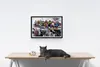 A framed painting of superheroes sitting on a city skyline is displayed on a white wall above a wooden table. A grey cat is lying on the table next to a small decorative object. Produktbild-Vorschau