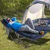 A man is relaxing on a camping cot next to a tent in a wooded area. Produktbild-Vorschau