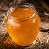 A close-up shot of a glass jar filled with golden honey, resting on a rustic wooden surface with dried straw scattered around. Anteprima dell’immagine prodotto