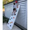 A young girl is climbing a white climbing wall attached to a white wooden fence. There is a red bell at the top of the wall. Voir les images du produit