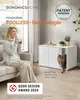 A woman sits on a couch holding a mug, next to a modern white cabinet with a cat emerging from it. The scene is set in a well-lit living room. Produktbild-Vorschau