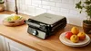 A waffle maker is shown on a kitchen counter next to a plate of apples and tomatoes, and a small bowl of food. Podgląd zdjęcia produktu