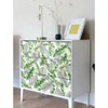 A white dresser with drawers decorated with a tropical leaf pattern, a gold lamp, and a framed picture on top. Produktbild-Vorschau