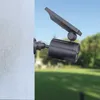 A solar-powered security light with a dummy camera is mounted on a textured wall, with a blurred background of a garden and sky. Produktbild-Vorschau