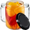 A close-up shot of three glass jars, one filled with red and yellow peppers, and a black lid placed in front of it. Two empty jars are visible behind the filled one. Podgląd zdjęcia produktu