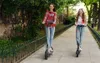 Two young women riding electric scooters down a paved path in a park-like setting. Produktbild-Vorschau