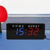A digital scoreboard for a table tennis game is placed on a blue table in front of a table tennis net. A red table tennis paddle is partially visible on the left. Náhled obrázku produktu