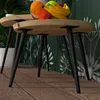 A close-up shot of two wooden side tables with black metal legs, placed on a wooden floor. A bowl of fruit sits on the table in the background, and a plant is visible on the left. The background features a green wallpaper with a white floral pattern. Produktbild-Vorschau