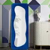 A tall, irregularly shaped mirror with a blue velvet frame stands against a wall with a geometric pattern. A white cabinet with a quilted texture is to the right of the mirror. Podgląd zdjęcia produktu