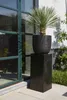 A black planter on a black pedestal is placed next to a large green-framed window. Produktbild-Vorschau