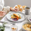 A person is eating breakfast with a set of tableware, including plates, cups, and croissants. Produktbild-Vorschau
