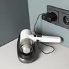 A white handheld vacuum cleaner is charging on its black base, plugged into a wall socket. Voir les images du produit