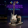 A miniature electric guitar and amplifier set on a stage with a neon "MUSIC FESTIVAL" sign in the background. Voir les images du produit