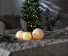 Three snowball-shaped candles with warm yellow light inside, placed on a textured gray tablecloth. A blurred Christmas tree with lights and a vase with fir branches are in the background. Produktbild-Vorschau