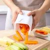 A person is using a white vegetable slicer to cut carrots over a wooden cutting board with other cut vegetables. Produktbild-Vorschau