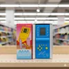 Two electronic handheld game consoles are displayed on a shelf in what appears to be a retail store. Náhled obrázku produktu