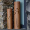 Two decorative cylindrical garden lights with a dandelion pattern are placed next to a stone wall and a blue door. Produktbild-Vorschau
