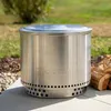 A stainless steel Solo Stove fire pit sits on a patio next to a pile of firewood. Produktbild-Vorschau