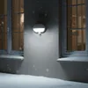 A white outdoor wall light with a motion sensor is mounted on a gray wall during a snowfall. Produktbild-Vorschau