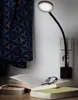 A black LED plug-in lamp is illuminating an open book and a pair of glasses on a table, next to a wall outlet and a patterned curtain. Produktbild-Vorschau