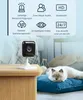 A white security camera is placed on a wooden table next to a fluffy cat lying on a blue cushion. The background shows a bedroom setting with a bed and curtains. Produktbild-Vorschau