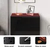 A black high-gloss nightstand with three drawers, topped with a red mat. Two framed pictures and a lamp are on the mat. Below the nightstand are three smaller images illustrating product features: water-proof, scratch-resistant, and easy to clean. Produktbild-Vorschau