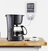 A coffee maker is plugged into a digital timer outlet on a wall, with a cup of coffee and coffee beans on a wooden surface in the foreground. Voir les images du produit