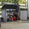 A man is organizing tools and equipment inside a large outdoor storage shed. Anteprima dell’immagine prodotto