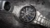A close-up shot of a men's wristwatch with a metal band, placed on a textured gray surface. A blurred object resembling a car gear shift is visible in the background. Produktbild-Vorschau