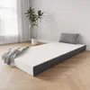 A foldable mattress is laid out on a wooden floor in a room with a large window and a plant. Produktbild-Vorschau