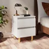 A white bedside table with two drawers and wooden legs, placed next to a bed. A lamp, a book, and a small plant are on top of the table. Náhled obrázku produktu