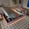 A pink upholstered bed with bedding and pillows, illuminated by LED lights underneath, set in a bedroom with a rug and artwork. Náhled obrázku produktu