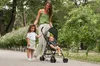 A woman is pushing a stroller with a baby in it, while a young girl walks beside them in a park. Produktbild-Vorschau