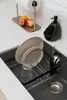 A dish rack with plates and cutlery placed in a kitchen sink, with fruit and a paper towel dispenser on the counter above. Produktbild-Vorschau